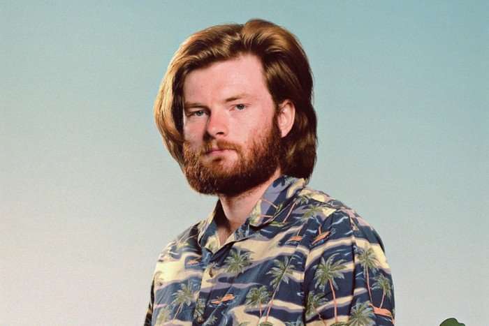 A man with ginger hair and beard poses for the camera, not smiling. He's wearing a green, cream and blue camo-style shirt