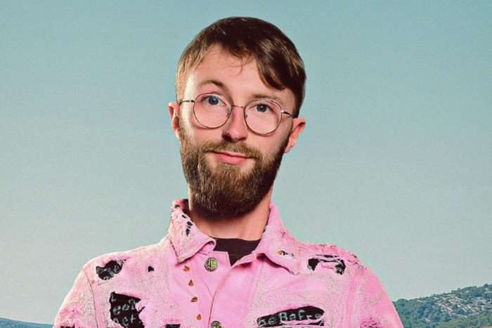 A slender man in a pink and black denim jacket poses for the camera. He has large round glasses and has short brown hair and a beard.