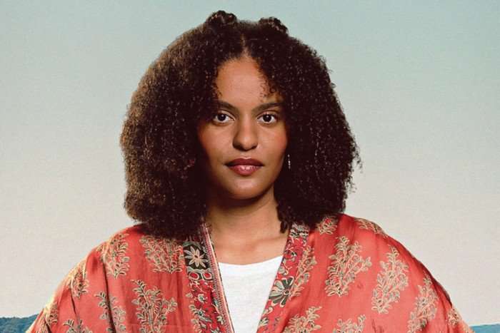 A woman with dark afro-style hair poses straight-faced for the camera. she's wearing a white t-shirt underneath a red and gold kimono