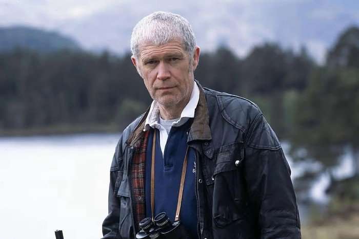 Alexander Morton in Monarch of the Glen, stood outside with binoculars around his neck and a lake behind him.