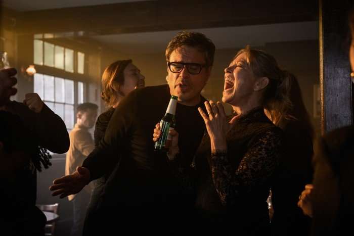 Alice and Steve at a party, holding bottles of beer and standing next to each other. Alice is laughing at something Steve has said.