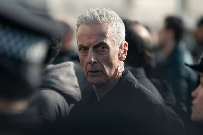 A close up of Peter Capaldi in a crowd of people looking to the right with a concerned expression.