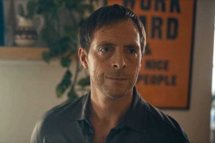 Stephen Campbell Moore in Criminal Record, wearing a shirt and stood inside, with an orange poster on the wall behind him.