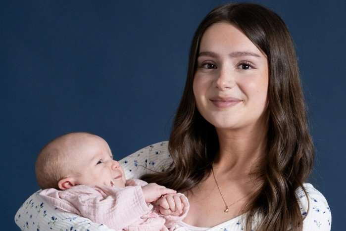 Sarah smiles at the camera holding baby Leyla, stood against a neutral deep blue background in key art for Emmerdale
