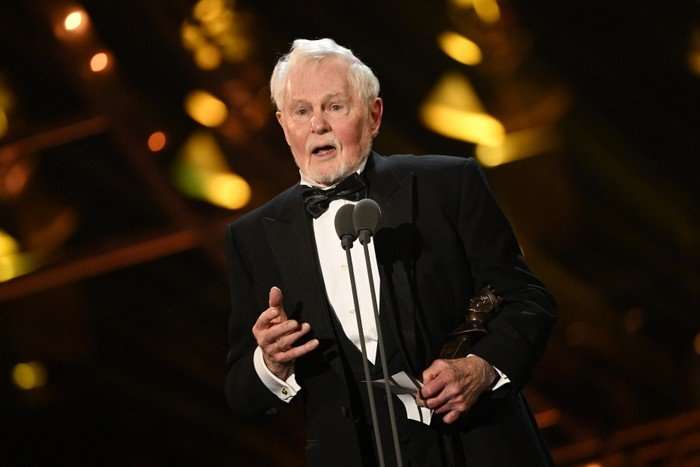 Derek Jacobi at a microphone in a tuxedo.
