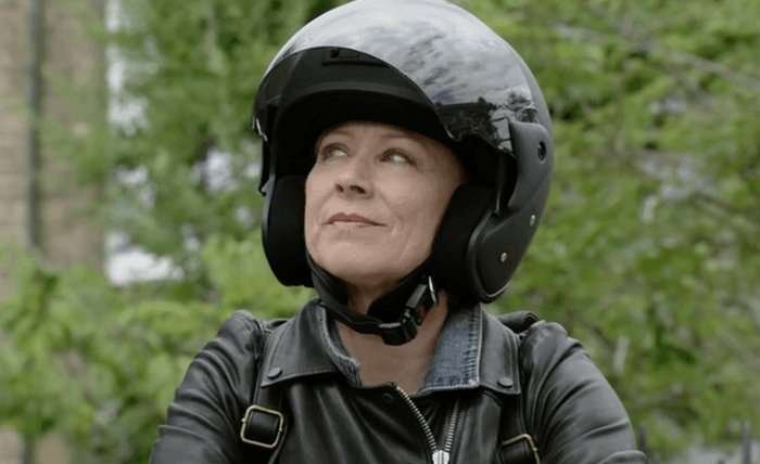 Lindsey Coulson as Carol Jackson smiling while sat on a motorcycle and wearing a helmet in EastEnders.