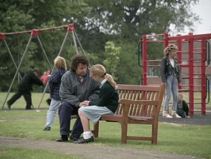 A long shot of Paul Bradley as Nigel Bates speaking with Gemma Bissix as Clare Bates on a park bench in a scene from EastEnders in the 1990s.