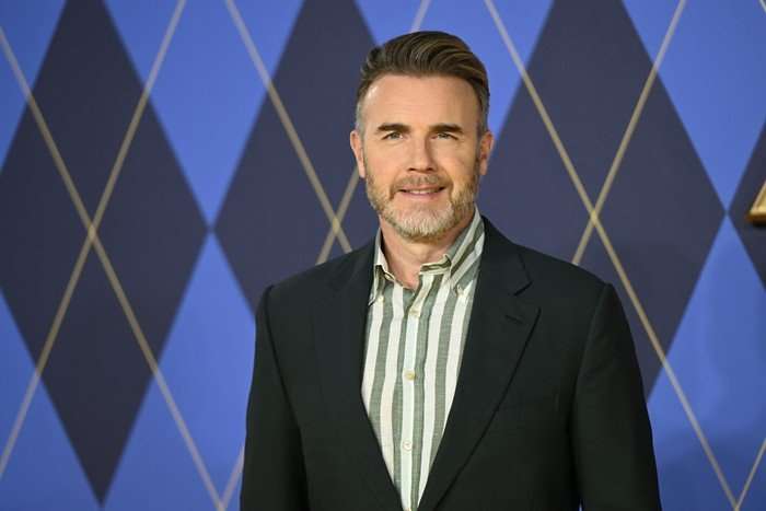 Gary Barlow wearing a striped shirt and blazer, smiling.