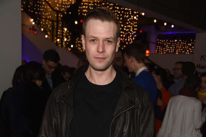 Sandy Batchelor, wearing a black t-shirt and leather jacket, stood at an event with people behind him.