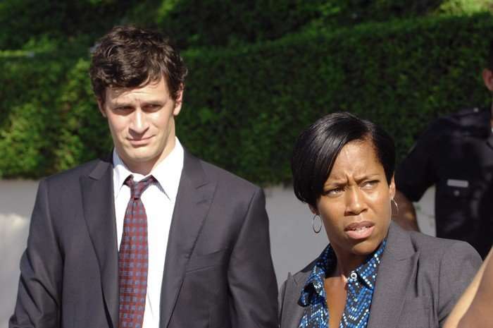A scene from Southland; Russell and Lydia wear work suits, while standing on the pavement on a sunny day. They look concerned and unimpressed at something to the right-hand side.