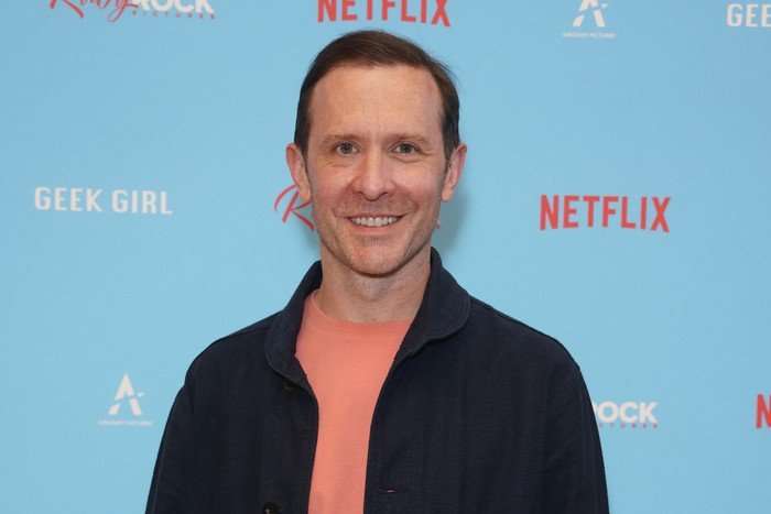 Tim Downie, wearing a pink t-shirt and blue jacket, stood in front of a light blue backdrop.