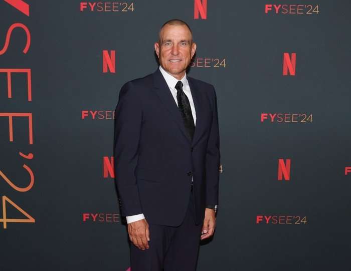 Vinnie Jones wearing a black suit and tie, smiling ahead.