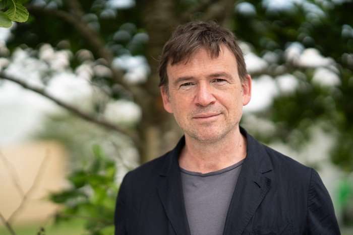 David Nicholls stands in a garden wearing a grey t-shirt with a black overshirt, he has short brown hair