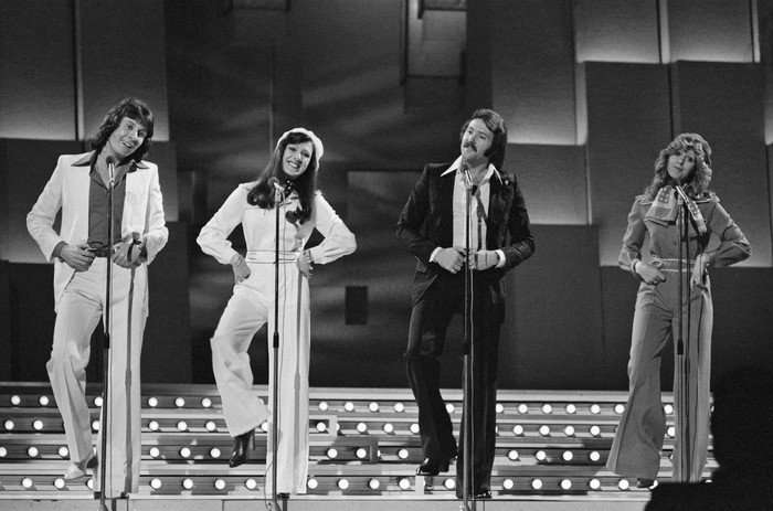 A black and white image of four people stood next to each other, smiling and singing.