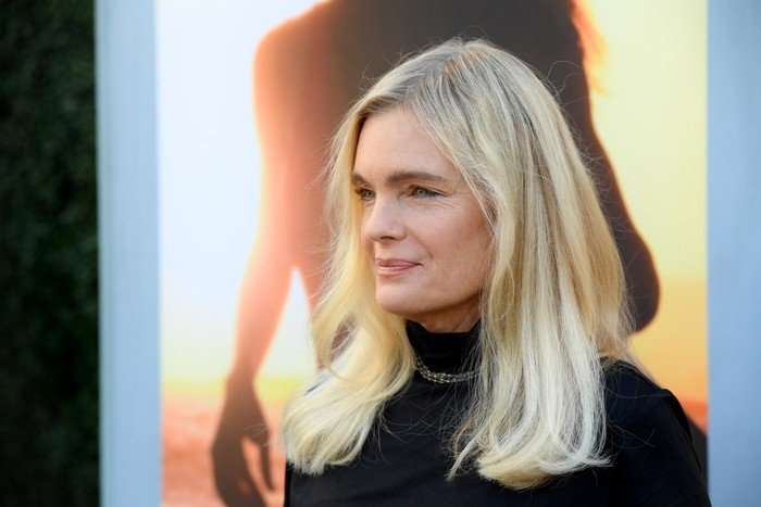 A blonde woman, Erika Eleniak, wears a black high neck jumper while posing for photographs