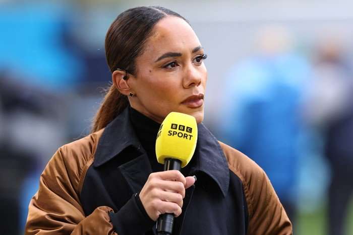 Alex Scott wearing a brown jacket, holding a yellow microphone and looking to the side.