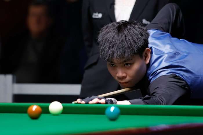 Snooker player He Guoqiang, in a black shirt and blue waistcoat, bends to take a shot on a snooker table.