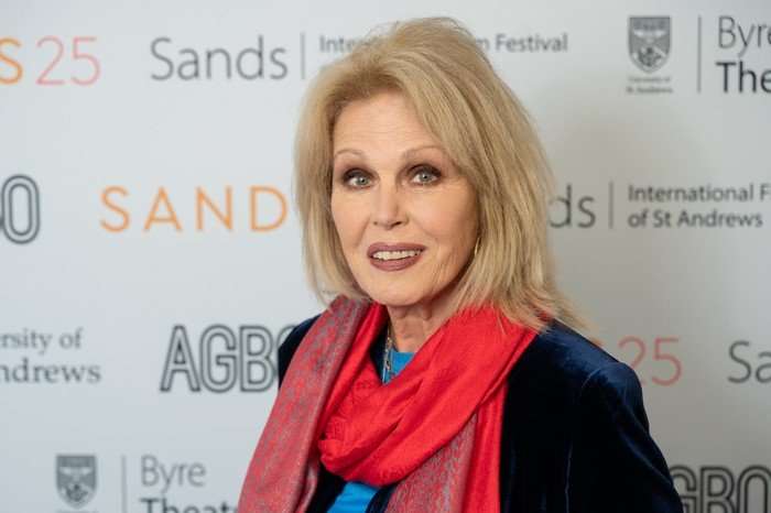 Joanna Lumley stands at a press call event. She is wearing a navy blue velvet blazer and a bright pink coloured scarf and has blonde hair