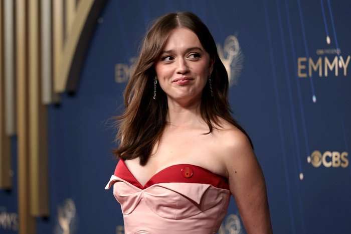 Aimee Lou Wood stood smiling as she looks to one side, wearing a pink and red dress.