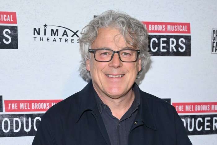 Alan Davies in black trousers, shirt and tie standing in front of a sign for The Producers play.