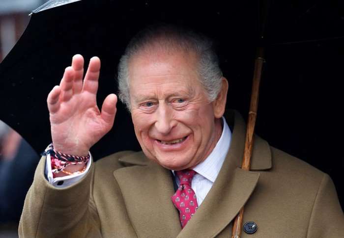 King Charles III stood holding an umbrella, waving to people in front of him.