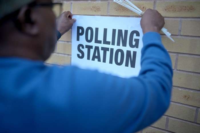 A man in a blue shirt fixing a sign saying Polling Station to a brick wall