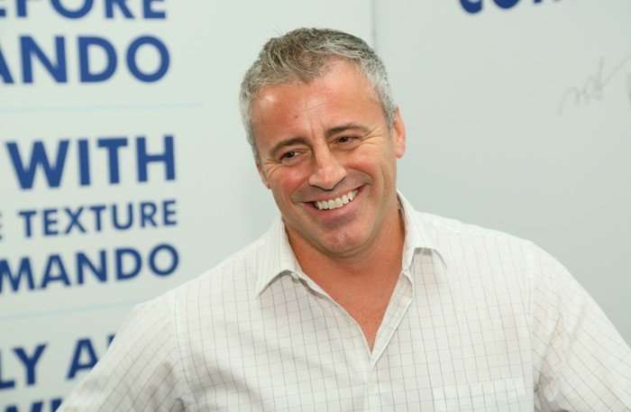 Matt LeBlanc is smiling in front of a white background in an open-collared white shirt with grey pinstripes.