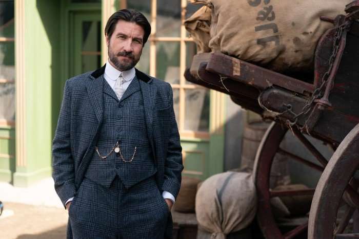 A smartly-dressed man in a three-piece navy suit stands in a street. He has a beard and longer hair, which he has brushed back away from his face