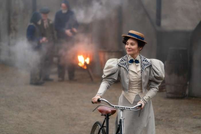 A woman in a full length pale blue dress, waistcoat, bolero and hat is walking while holding a bicycle. There is a fire behind her