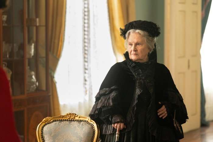 An older woman, dressed in 19th century black funeral attire, looks stern as she stands by a window