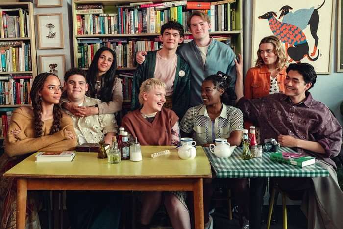 The main characters of Heartstopper sat and stood around a table at a cafe, laughing and smiling together