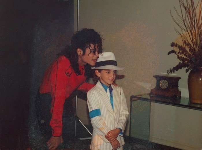 Michael Jackson wearing a red shirt and black trousers, crouched down next to a young boy wearing a white suit and hat.