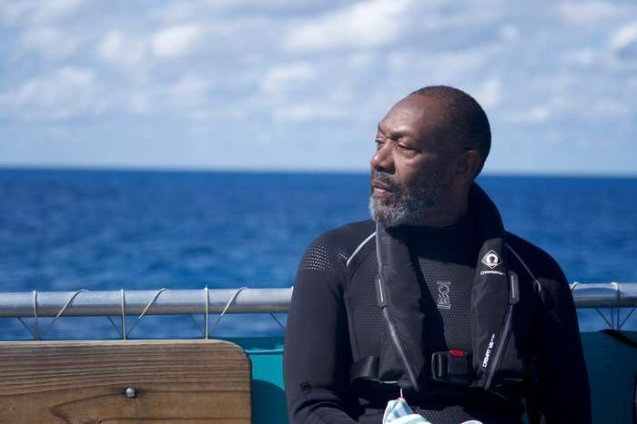 Sir Lenny Henry sitting on a boat in a wetsuit, looking out at the sea.