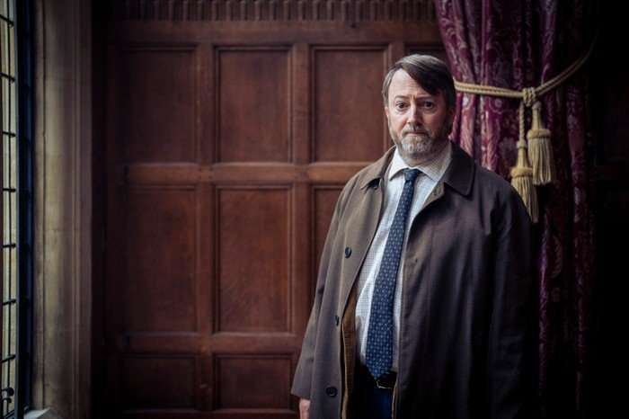 David Mitchell as John ‘Ludwig’ Taylor in Ludwig, stood against a wall boarded with wood.