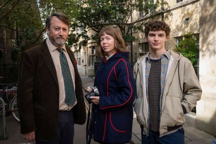 David Mitchell as John ‘Ludwig’ Taylor, Anna Maxwell Martin as Lucy Betts-Taylor and Dylan Hughes as Henry Betts-Taylor in Ludwig. They are stood together in a street and Martin is holding a camera.