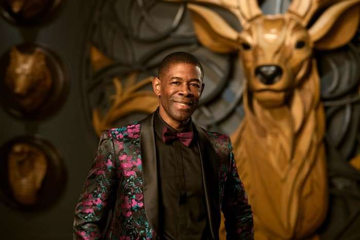 A man smiling ahead, wearing a black and purple coloured tuxedo, smiling with his hands on his hips.