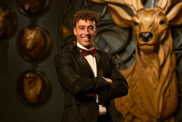 A man smiling ahead with his arms folded in front of his chest. He is wearing a black tuxedo with a red bowtie.