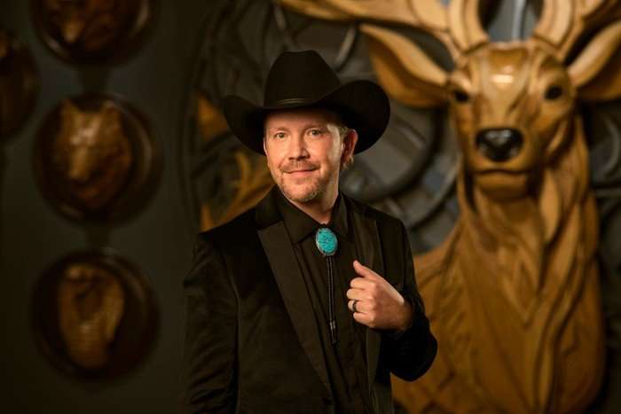 A man wearing an all black suit paired with a black cowboy hat, smiling ahead, with one hand on his lapel.
