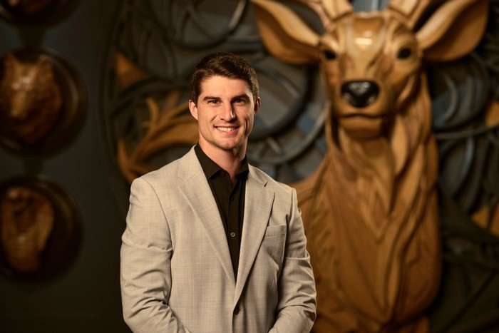 A man stood smiling ahead, wearing a black shirt and grey blazer.