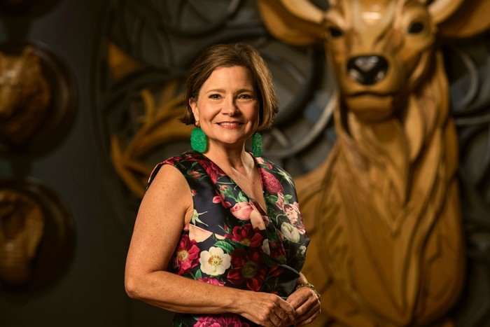 A woman smiling ahead with her body turned slightly to one side. She is wearing a floral dress and bright green earrings.