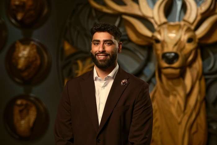 A man smiling ahead as he poses for a photo. He is wearing a white shirt and a dark red blazer.