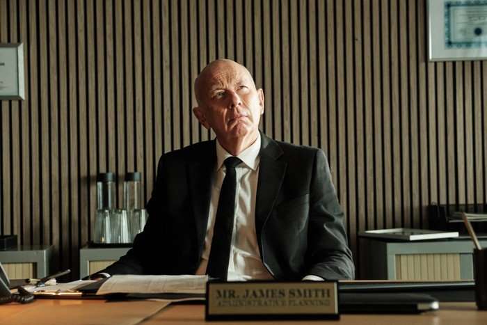 A bald man in a black suit and tie sits at a desk, looking upward thoughtfully. A nameplate reading &ldquo;Mr James Smith&rdquo; is in front of him, with papers and office items on the desk and a wood-paneled wall behind.