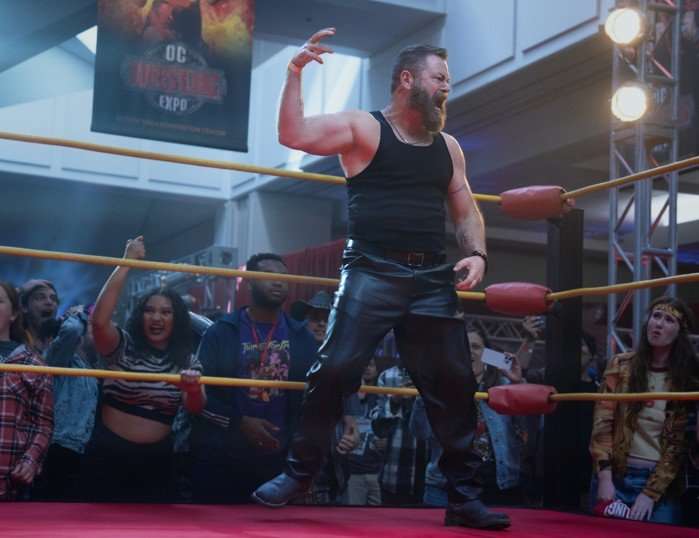 Nick Offerman Nick Offerman stands in a wrestling ring raising one hand to the air in celebration