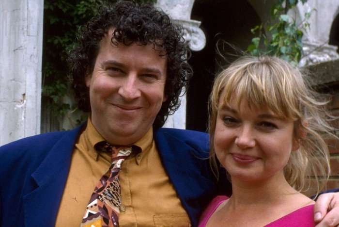 Nigel, in a blue blazer and yellow shirt and patterned tie, stands beside Debbie, who is wearing a pink blouse, in a shot from EastEnders in the 90s