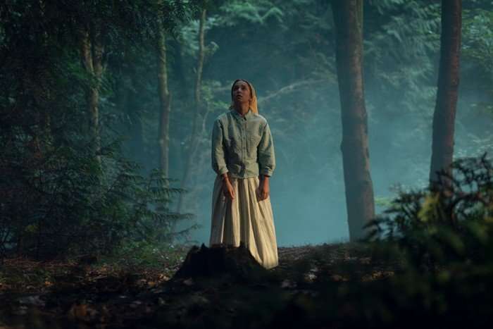 Molly Windsor in Unchosen, wearing a blue top and white dress and stood in a forest.
