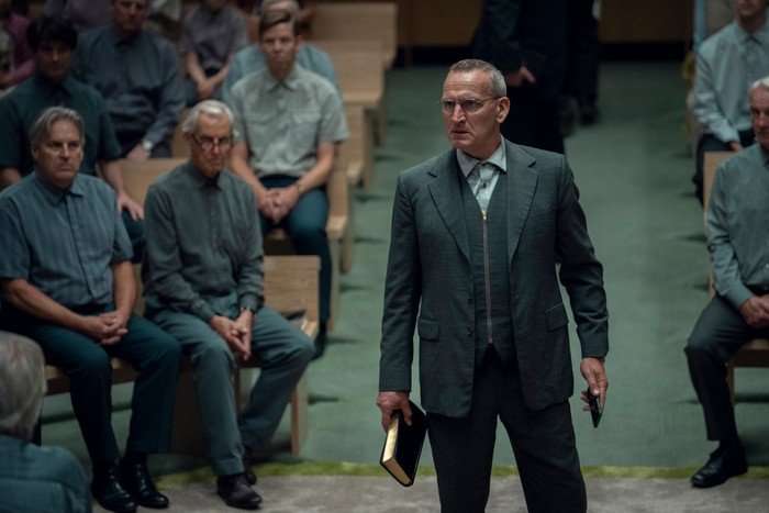 Christopher Eccleston stars in Unchosen. He is standing in the centre of a hall, where a service is taking place, with uniformed attendees sat around him.