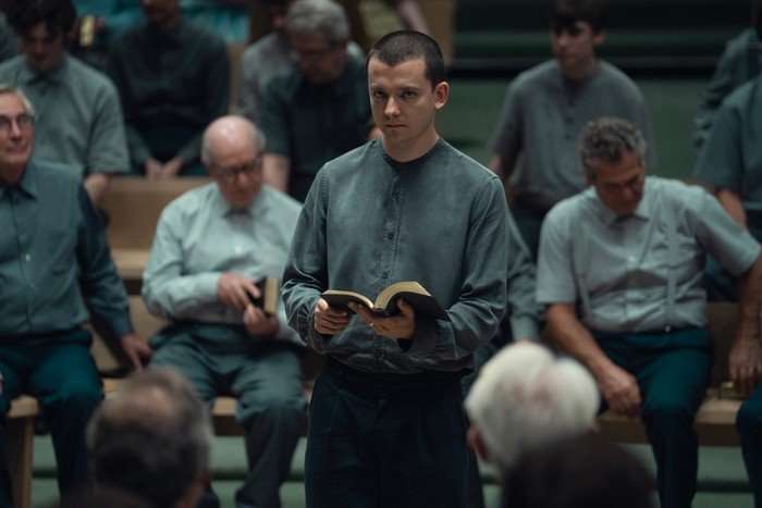 Asa Butterfield in Unchosen, wearing grey and holding a book, with men sat all around him.