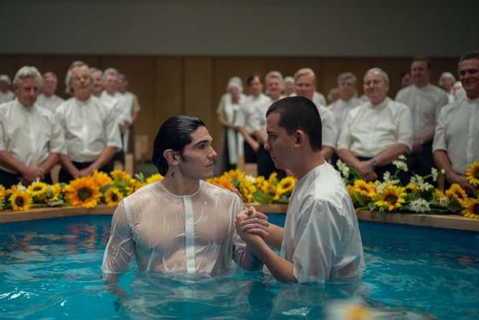 Asa Butterfield and Fra Fee in Unchosen. They are both wearing white shirts and stood together in a pool. A group of men in white shirts are stood in the background, watching them.