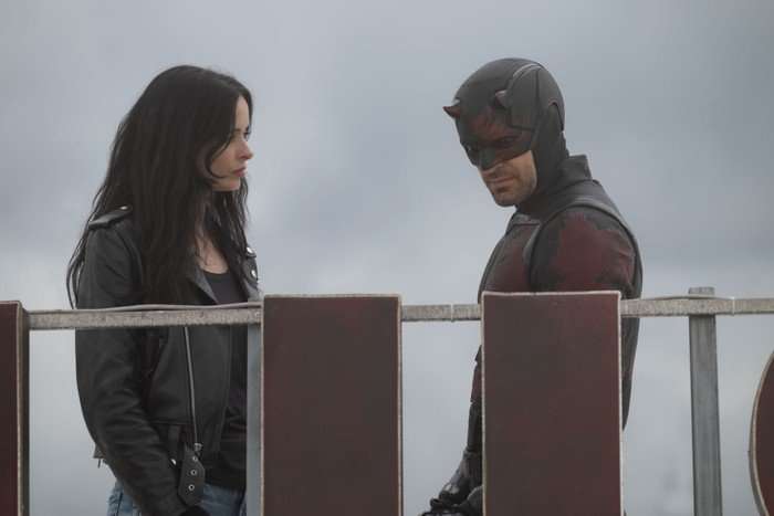 Krysten Ritter as Jessica Jones and Charlie Cox as Matt Murdock / Daredevil in Daredevil: Born Again, stood together against a grey sky. He is wearing his daredevil costume and she is wearing a black leather jacket and top.