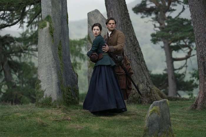 Hermione Corfield and Jeremy Irvine in Outlander: Blood of My Blood season 2 standing in the middle of a forest, surrounded by stones but turning back and looking scared.